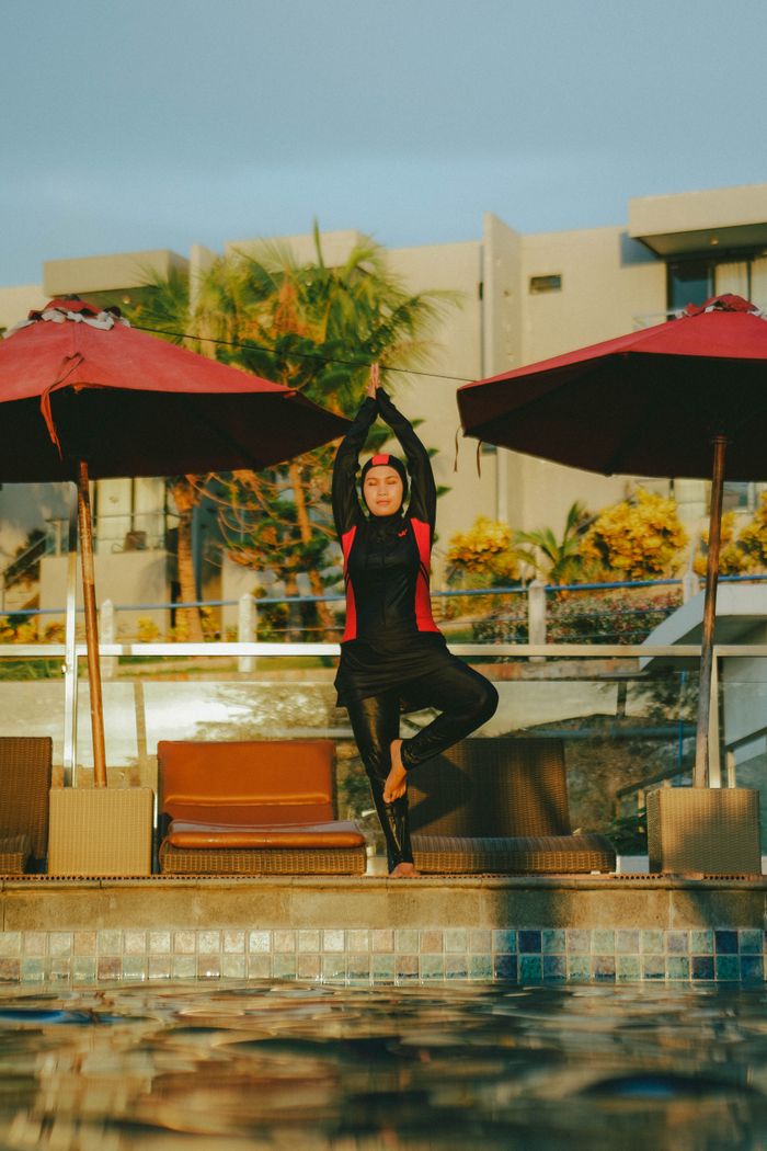 A woman in swimwear performs yoga by a pool, surrounded by umbrellas and deckchairs.